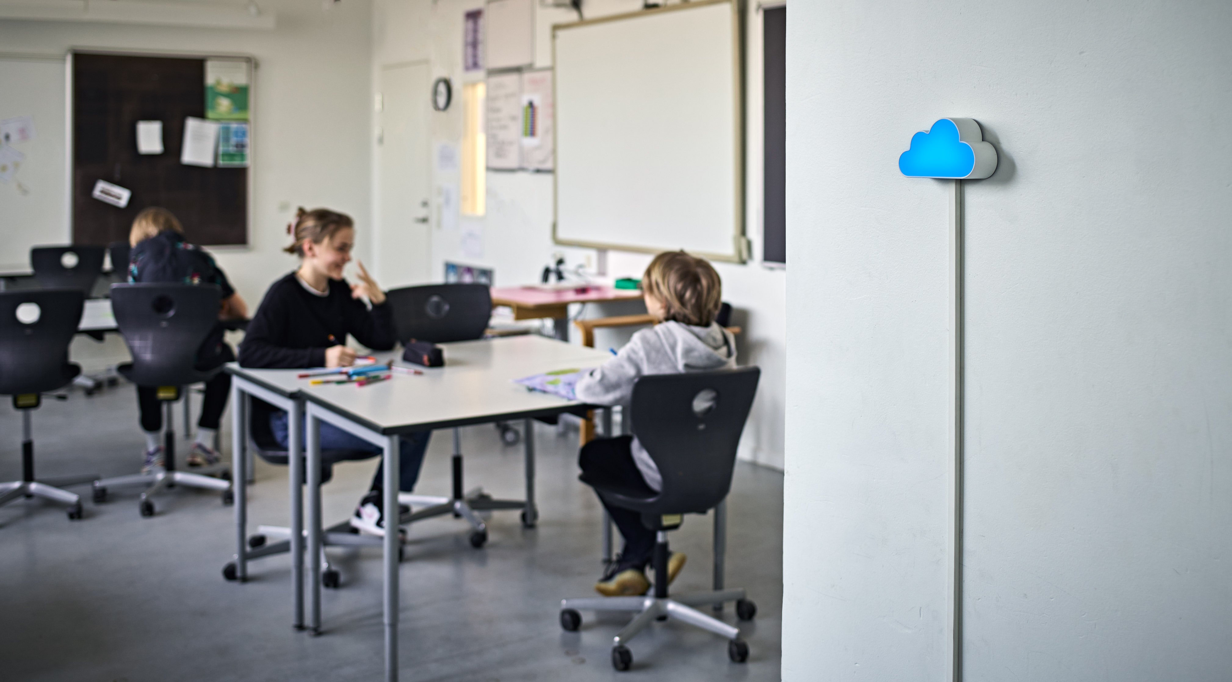 CO2-måleren SKYEN lyser blåt på væggen i et skolelokale med arbejdende børn